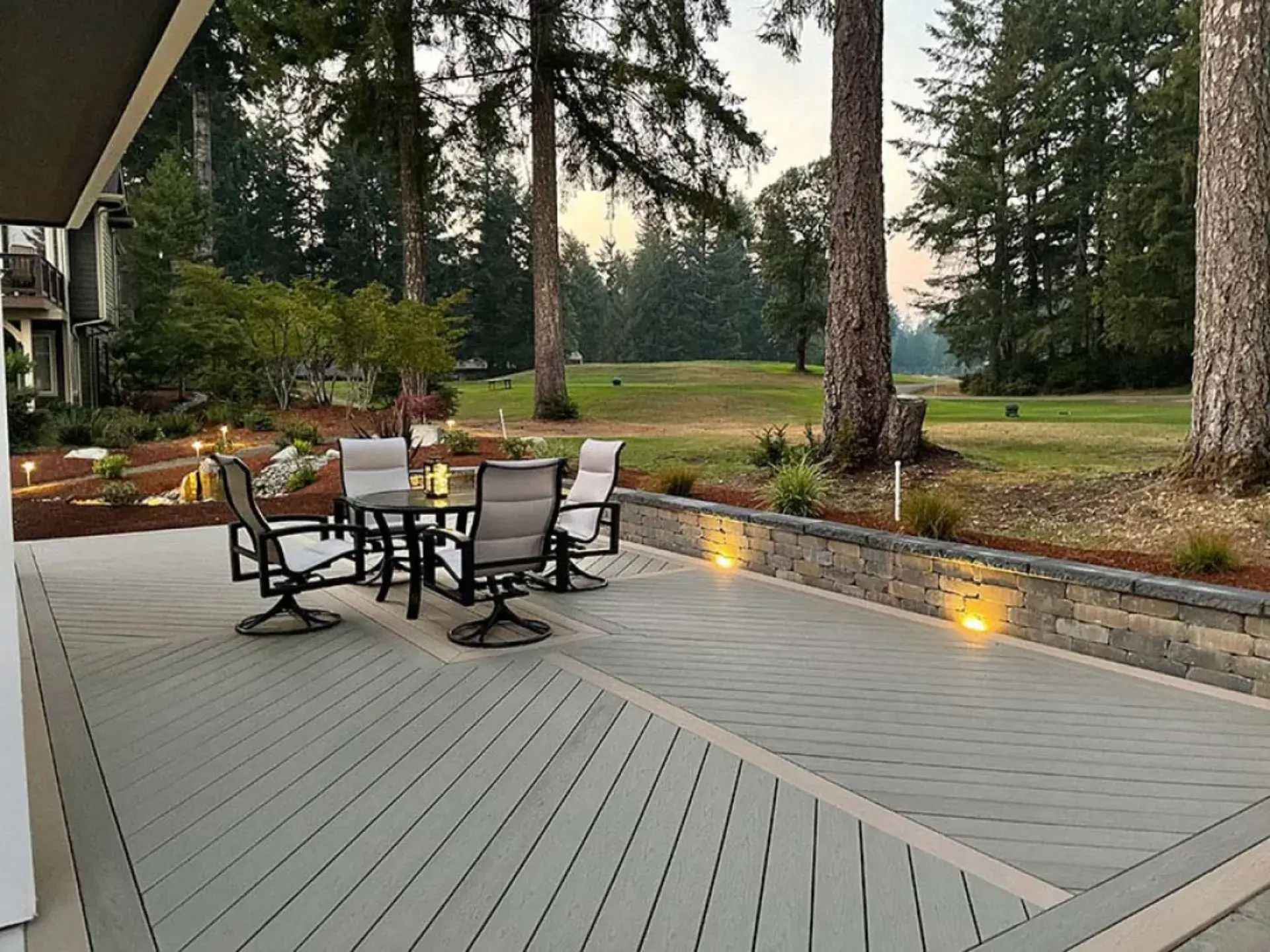 Patio with outdoor table and chairs on a gray deck, overlooking a landscaped backyard with tall trees and warm lights