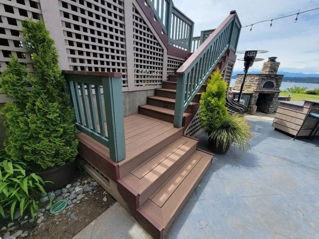 Outdoor wooden staircase with brown steps and green railings beside a patio and stone fireplace