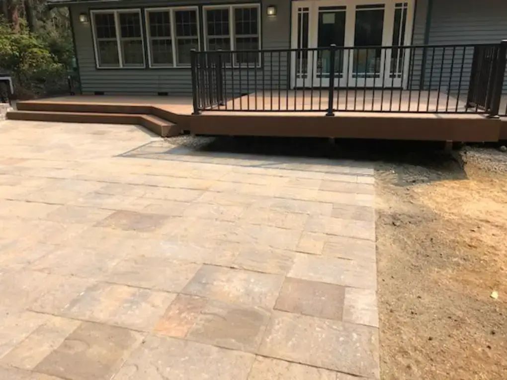 Patio with stone pavers, raised wooden deck, and black railing in front of a house