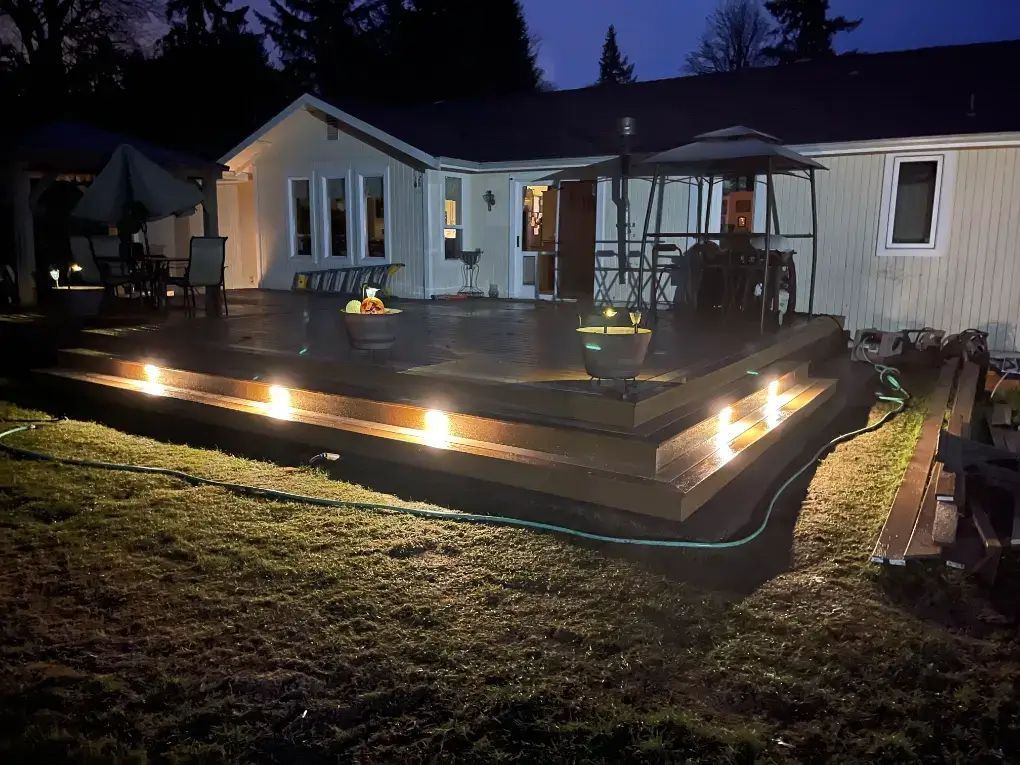 Backyard deck at dusk with string lights, patio chairs, and a fire pit beside a house