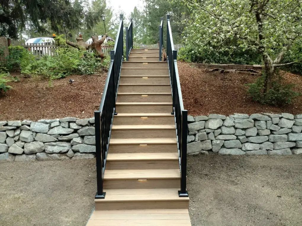 Outdoor staircase with black railings leading up a landscaped slope, bordered by stone retaining walls.