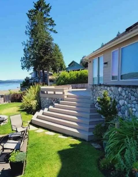 Backyard patio with stone steps, lawn, potted chairs, and a house by the water
