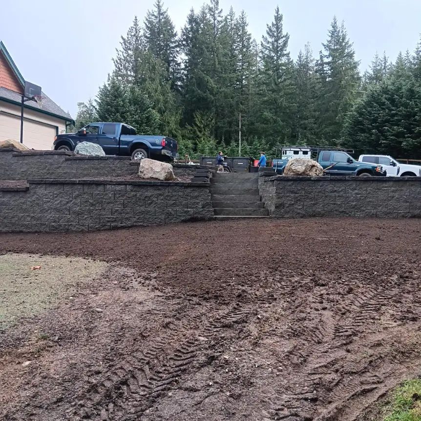 Construction site with muddy foreground, stone retaining wall, steps, and parked trucks and cars by evergreens.