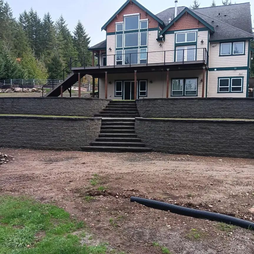 Large house with stone retaining wall and central stairs, surrounded by trees and a dirt yard.