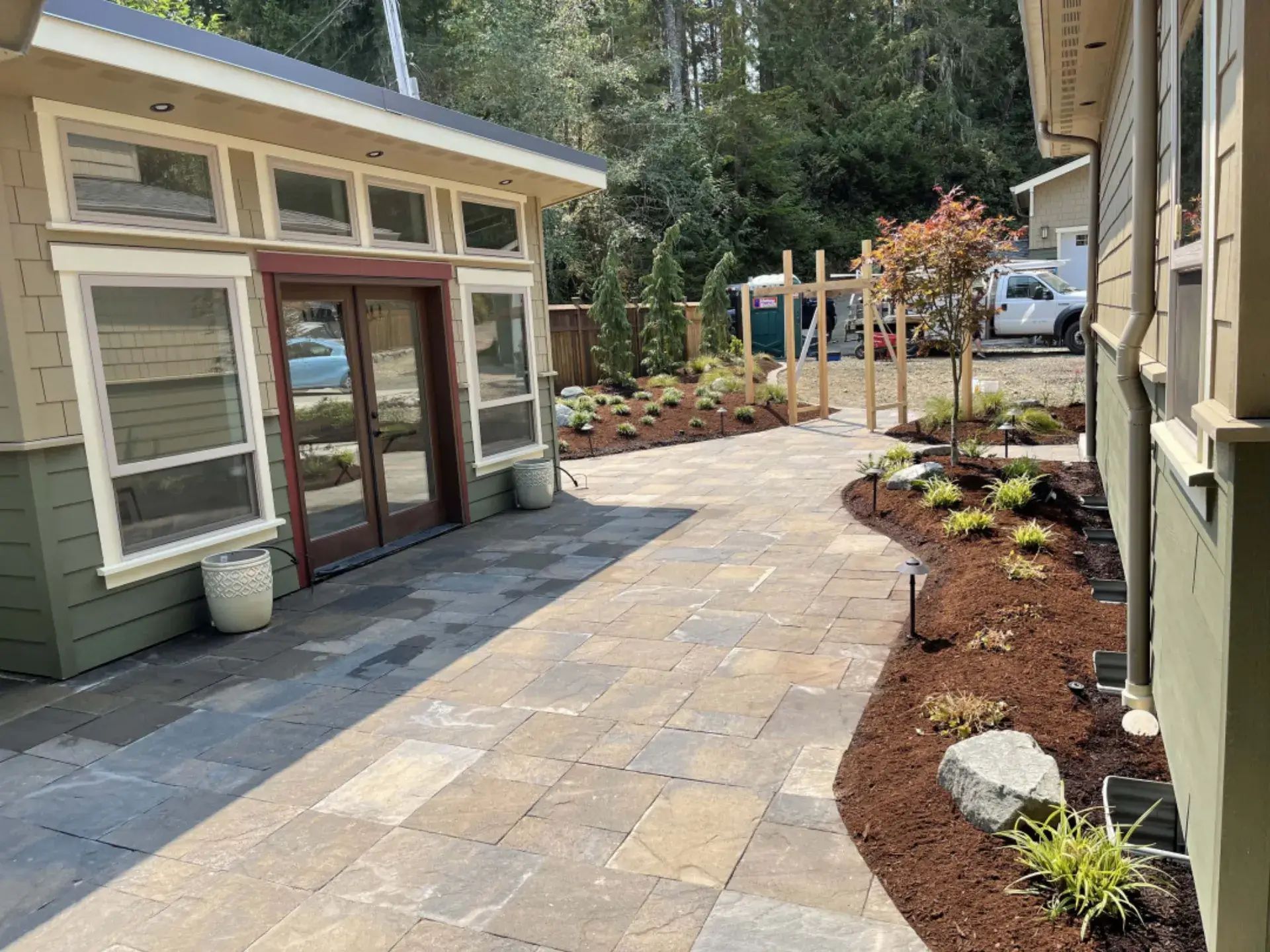 Stone patio walkway between beige buildings with garden beds and trees, leading to a driveway and parked car.