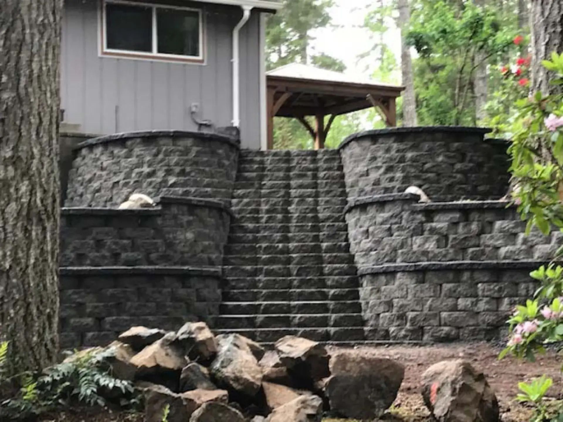 Stone stairway between retaining walls leading to a house, with rocks scattered at the base and greenery nearby.