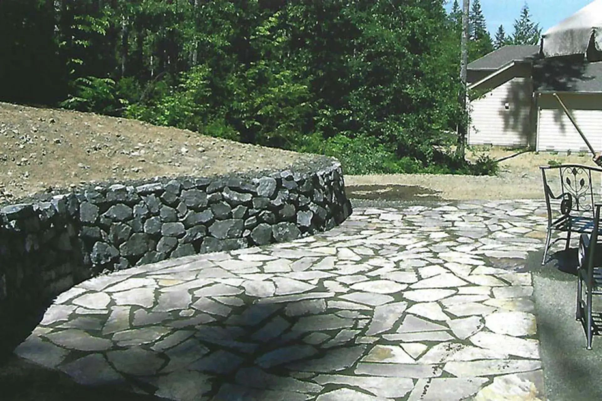 Stone patio and retaining wall beside a building, with trees and bright sunlight in the background.