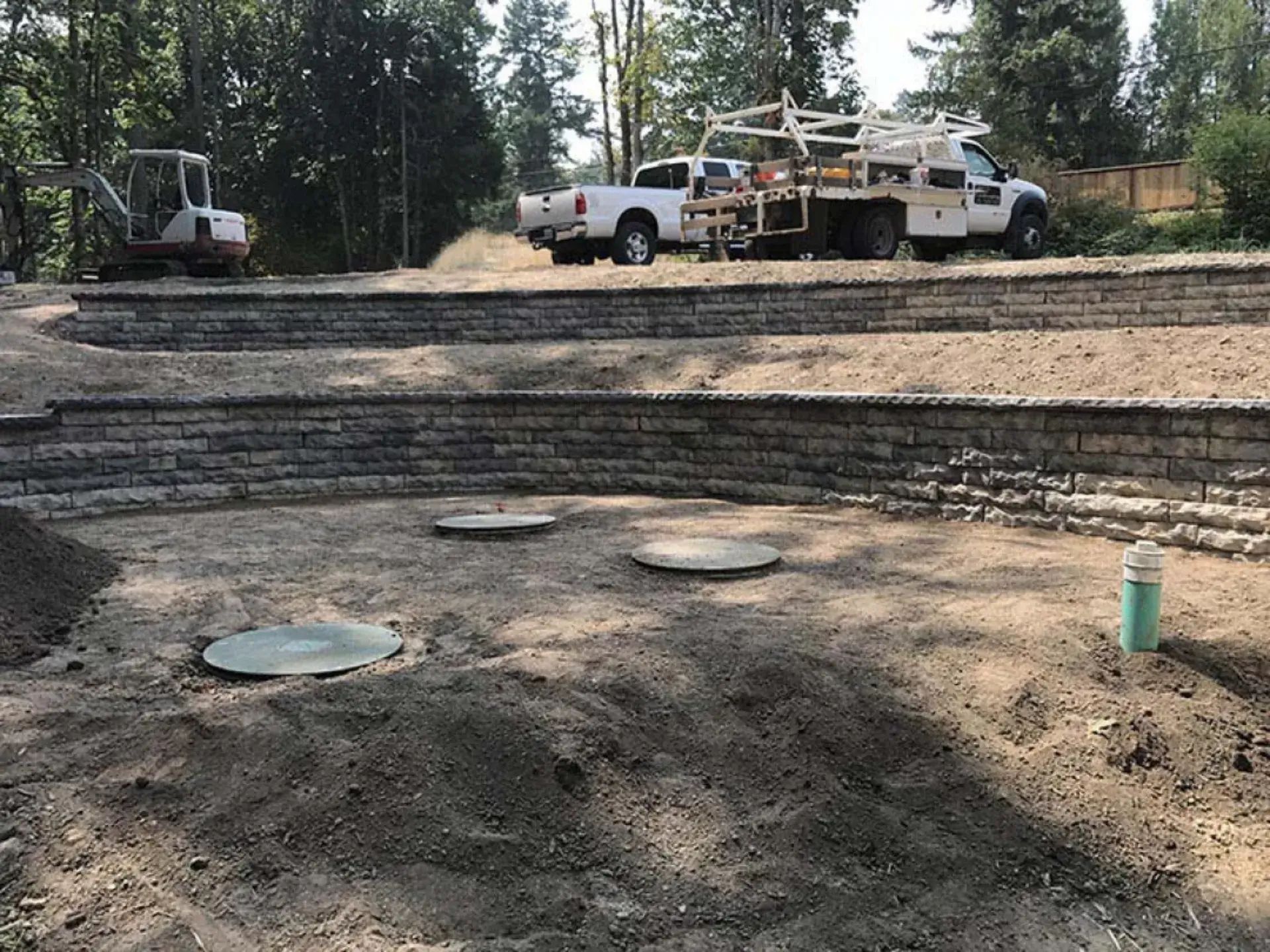 Construction site with trucks, gravel terraces, utility covers, and trees in the background