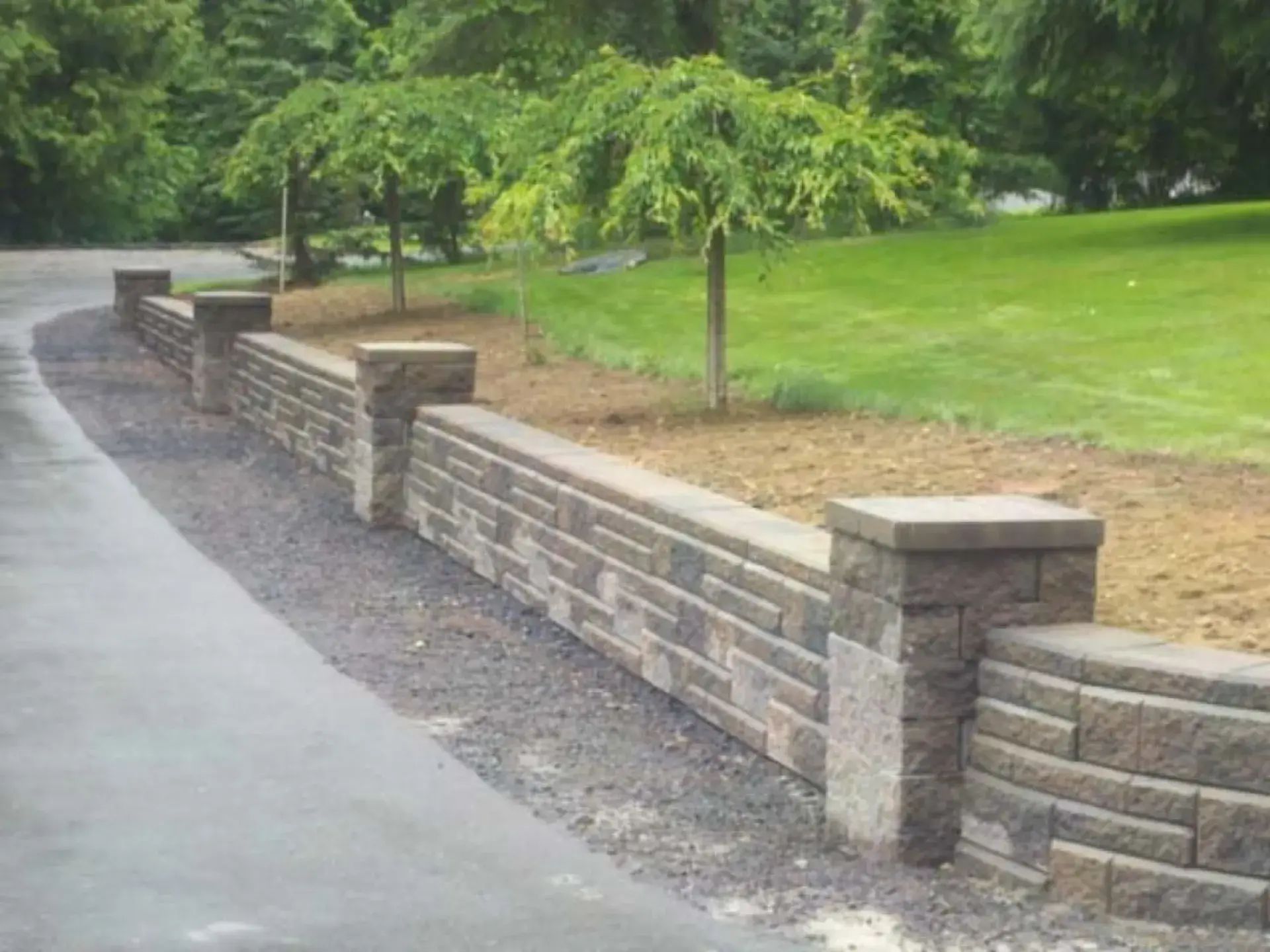 Curved stone retaining wall beside a paved path and green lawn with trees in a park