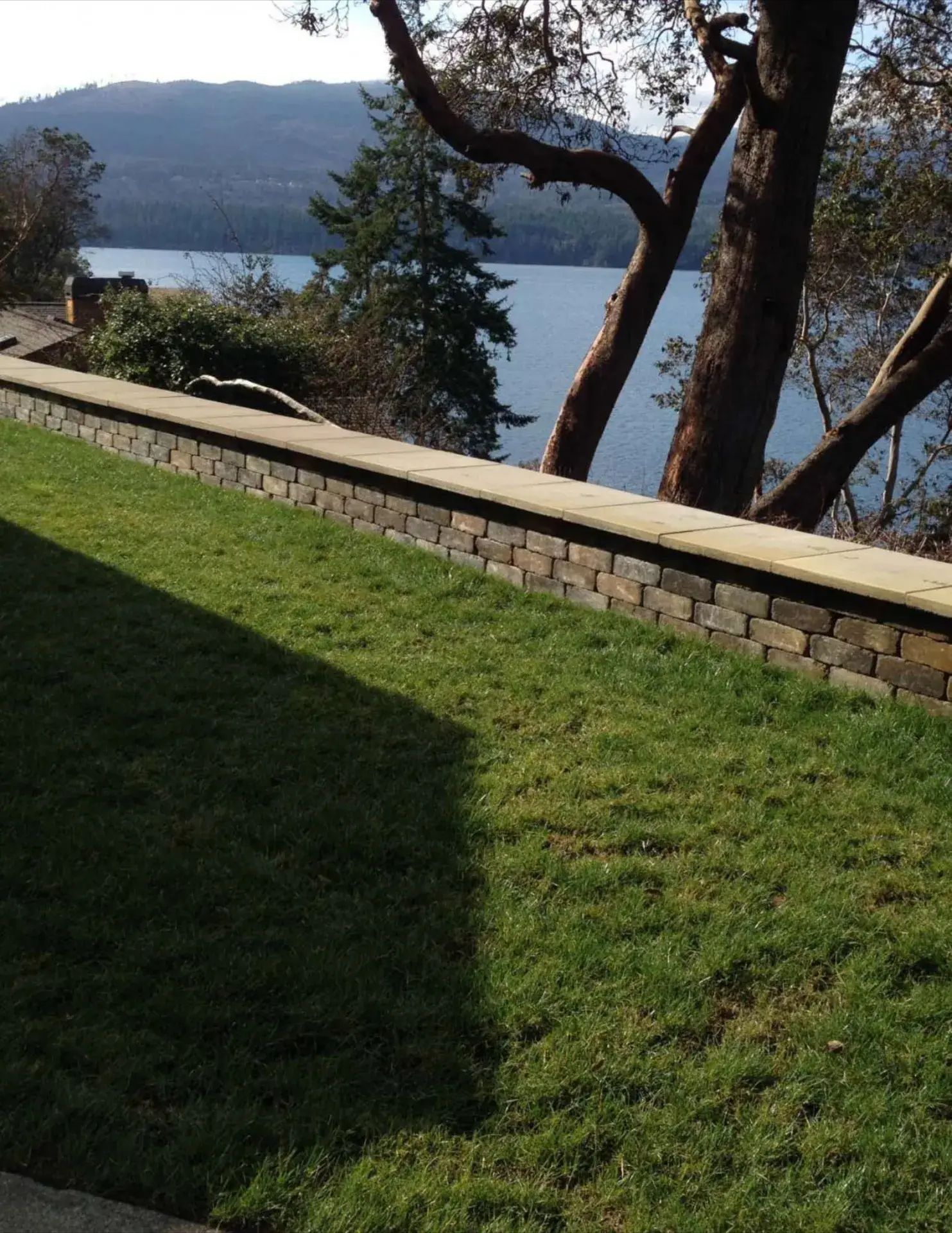 Sunlit grassy yard beside a stone wall, overlooking a lake and distant hills through trees