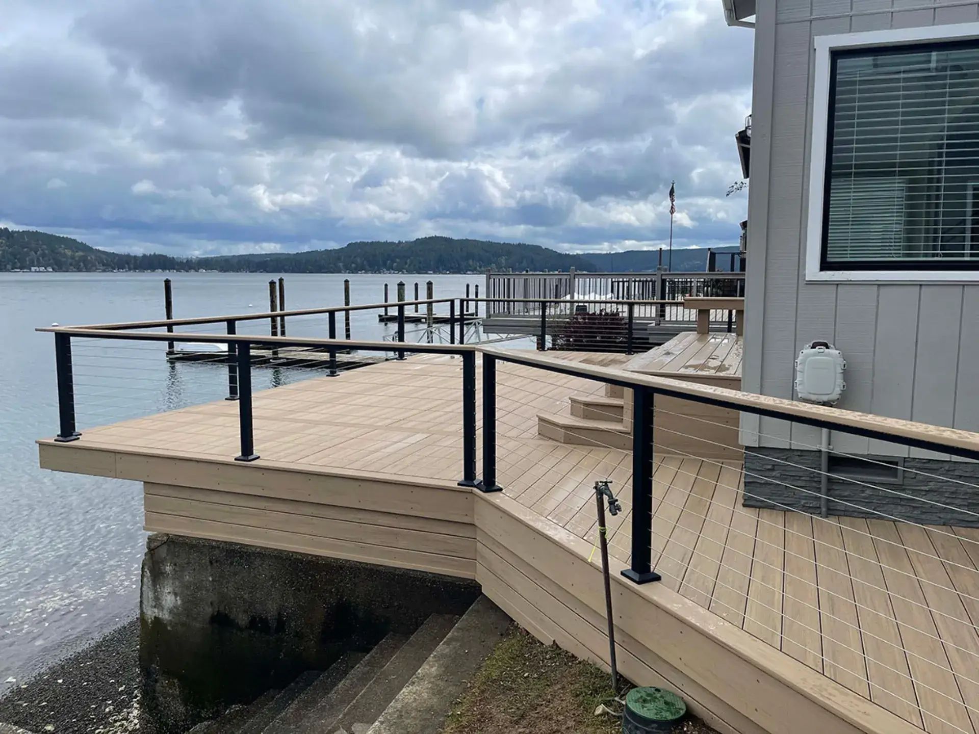 Wooden deck beside a waterfront house, with black railings and cloudy sky over calm water.