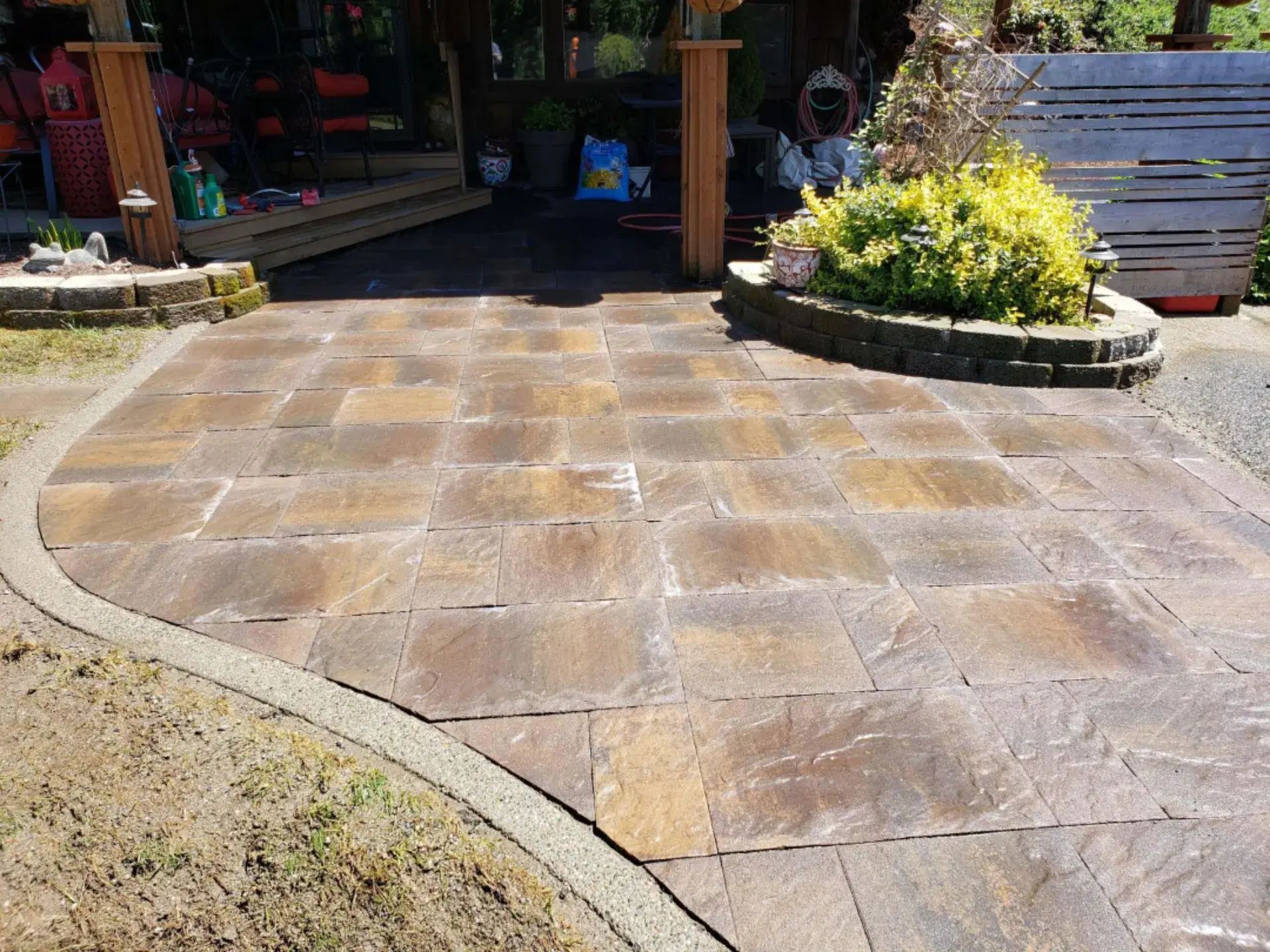 Sunlit stone patio with a curved lawn edge and potted plants by a garden entrance