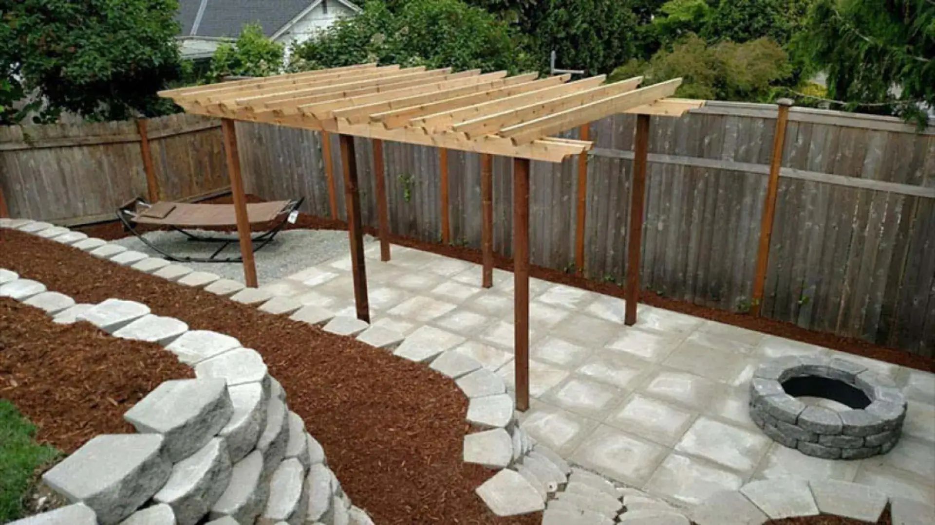 Backyard patio with wooden pergola, stone pavers, mulch beds, and a small fire pit
