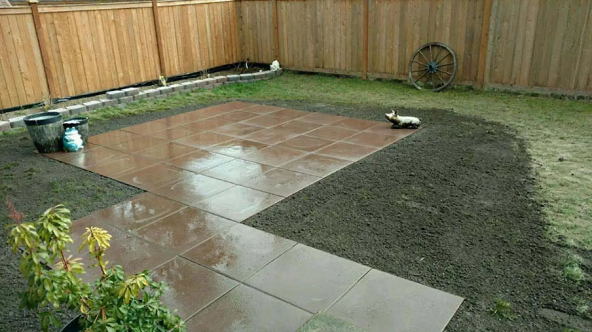 Backyard patio with wet tile and a dark, freshly leveled soil bed beside a wooden fence.