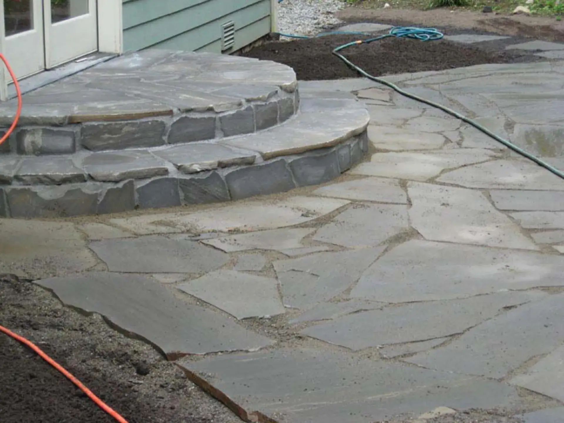 Stone patio with curved concrete steps beside a house and a dirt area under construction