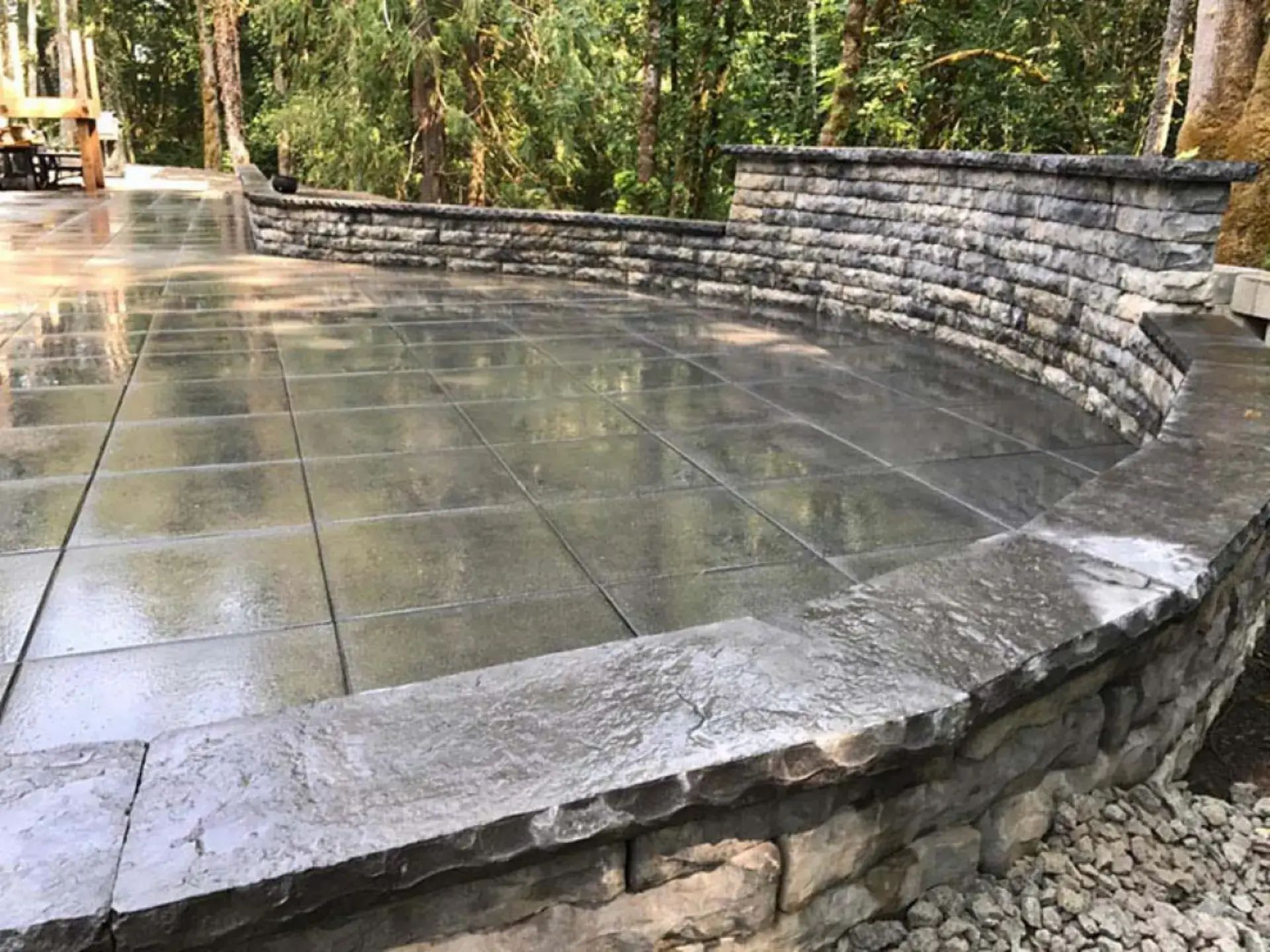 Curved stone patio with wet concrete pavers and a low stone wall in a wooded park setting