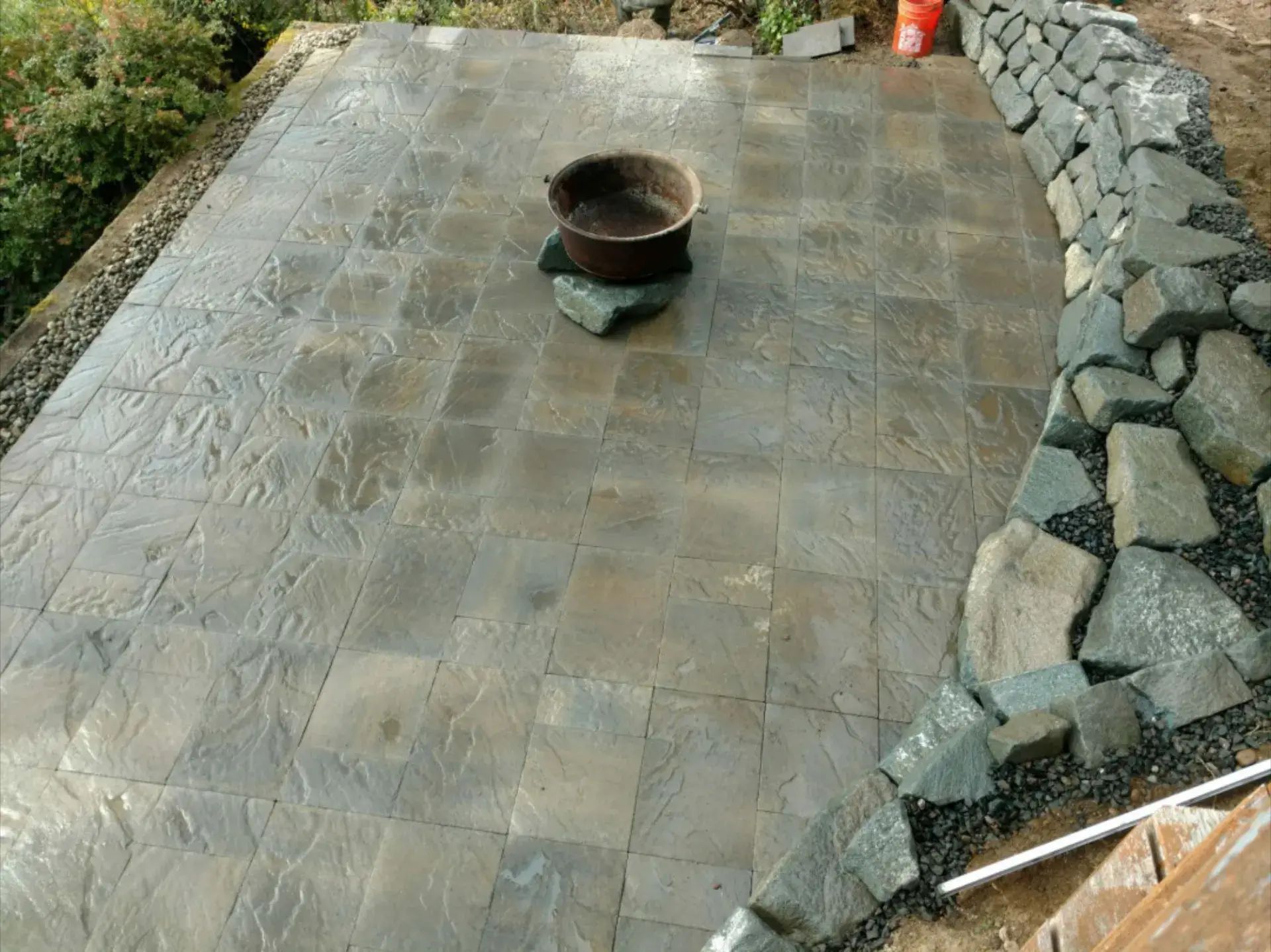 Stone patio under construction with a central fire pit and stacked rocks along one edge.
