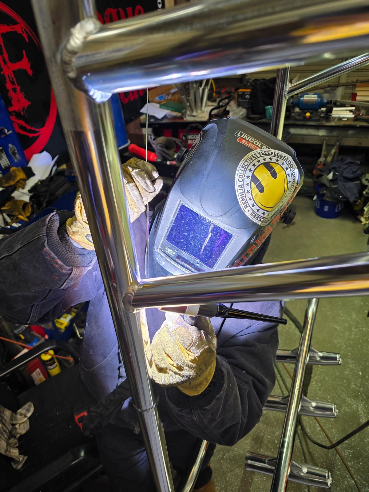 Welder wearing a helmet, gloves, and dark clothing welds metal framing in a shop.