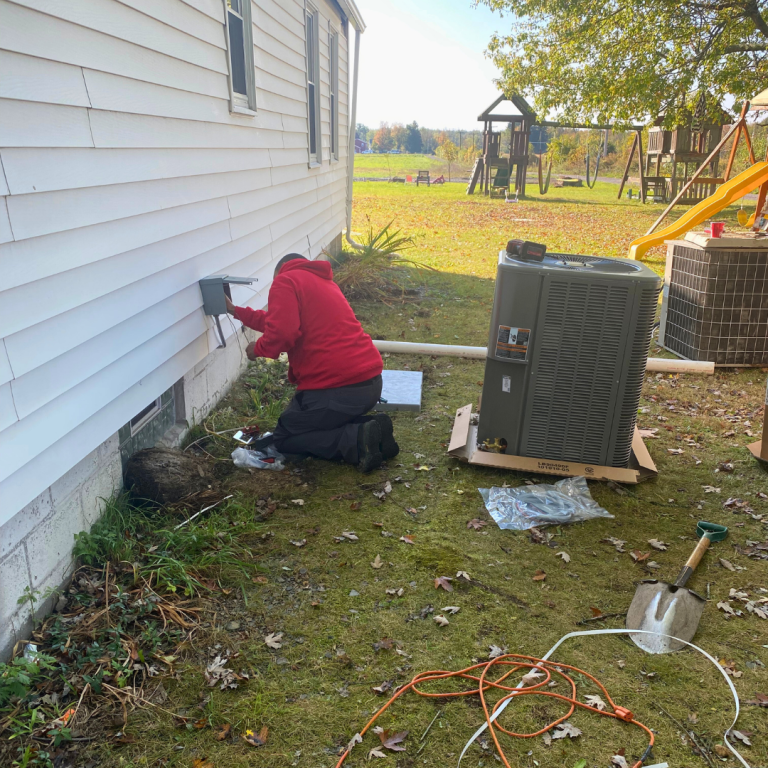 A person in a red hoodie kneels by a house, installing an outdoor electrical disconnect for a new air conditioning unit.