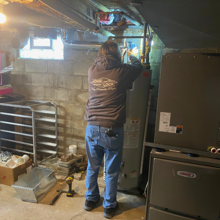 A person wearing a hooded sweatshirt works on water heater pipes in a basement next to a large furnace unit.