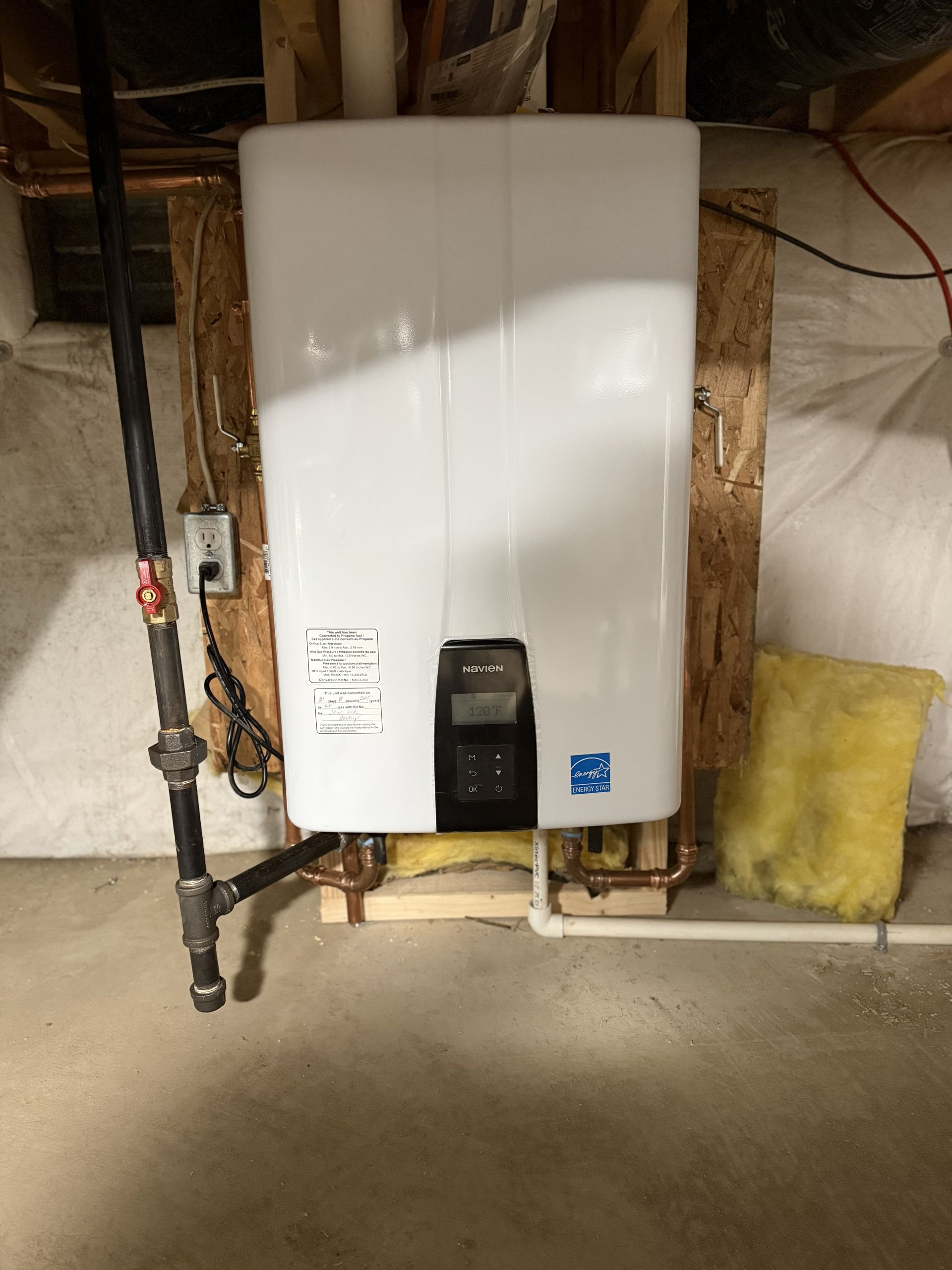 White wall-mounted water heater with pipes in an unfinished basement; yellow insulation on the right.