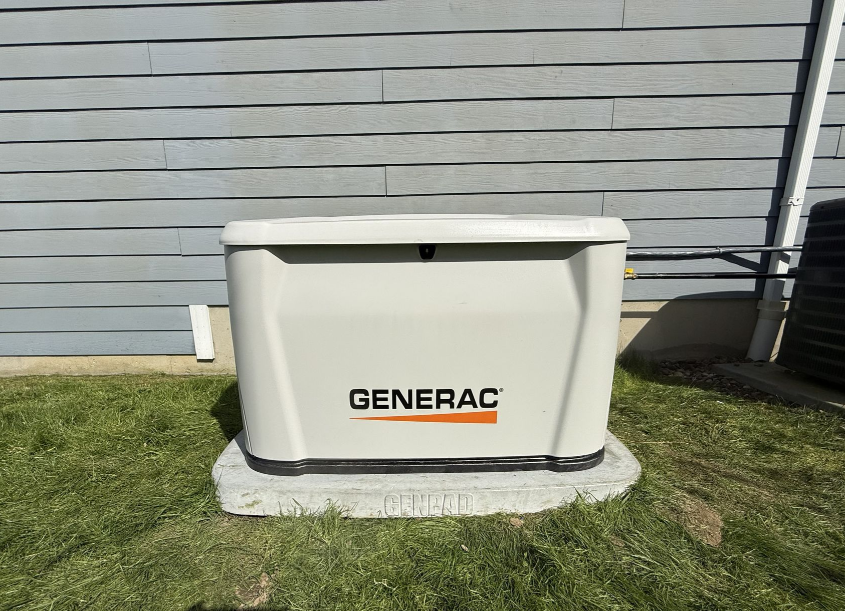 A beige home standby generator sits on a gravel pad next to the white siding of a house.