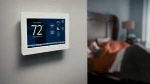 A white smart thermostat mounted on a wall displays 72 degrees, with a blurred bedroom visible in the background.