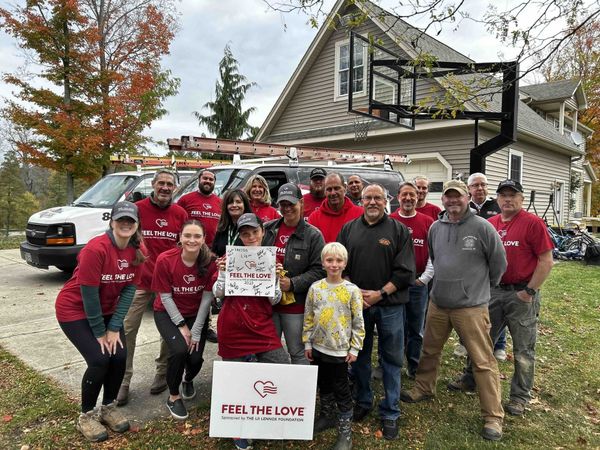 A group poses in front of a house and a work van, holding a