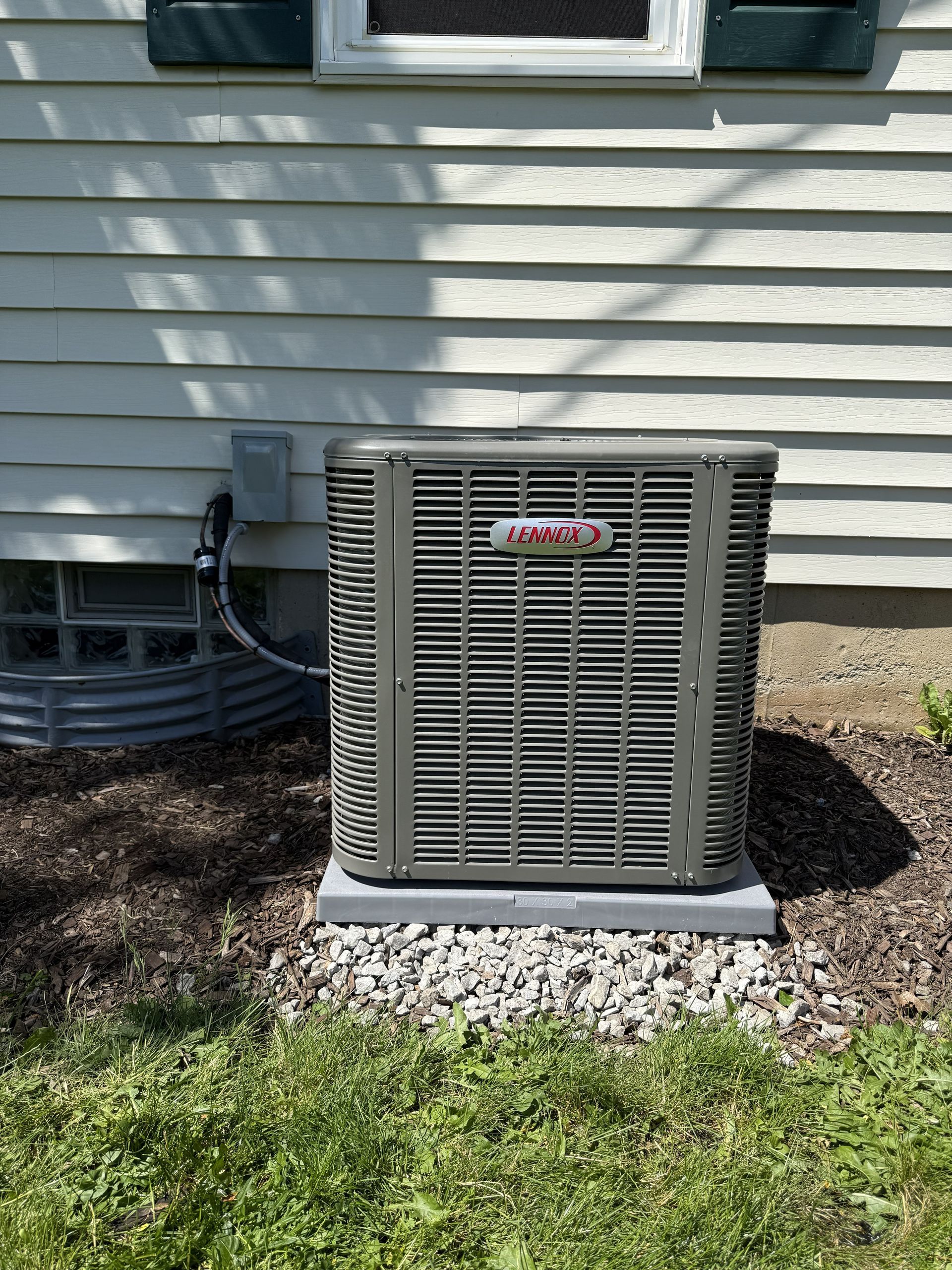 A gray, modern heat pump unit sits outside a white building, framed by small evergreen trees on a gravel walkway.