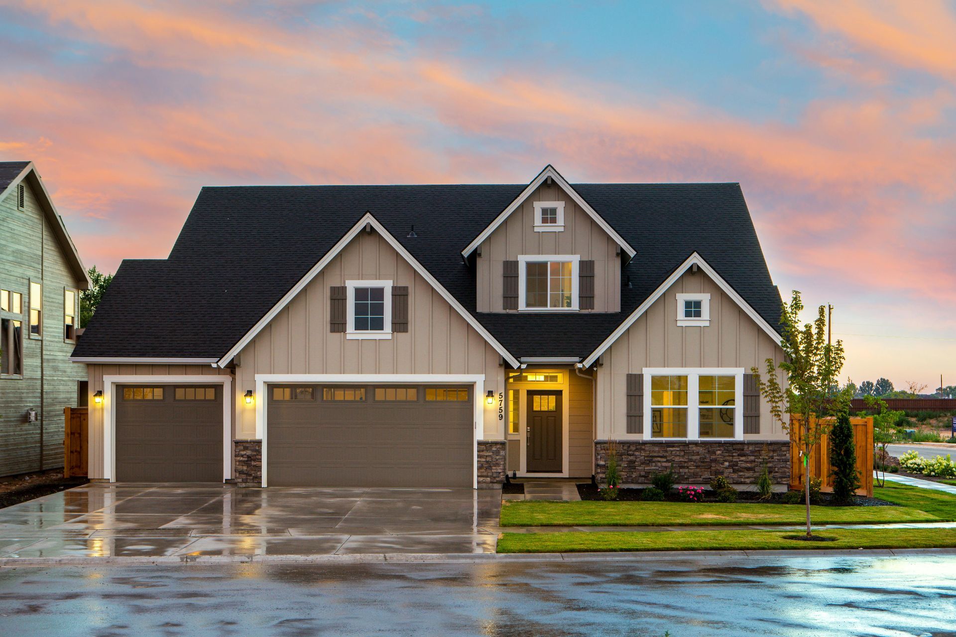 A two-story suburban house with beige siding, dark stone accents, a three-car garage, and a sunset sky background.