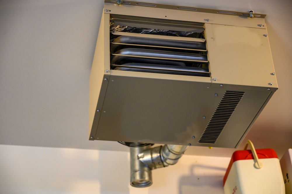 A beige ceiling-mounted gas unit heater with a metallic flue pipe, installed indoors near a white container.