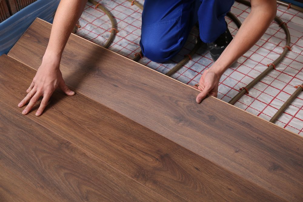 A person in blue work pants installs dark wood laminate flooring over an underfloor heating system.