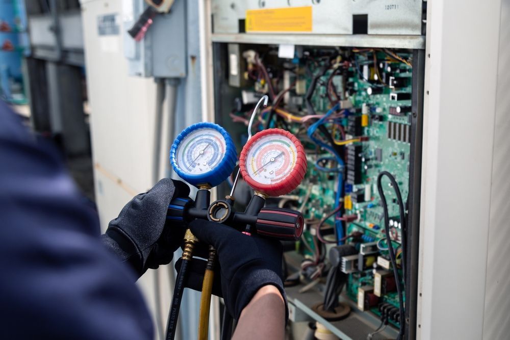 A technician in gloves holds a manifold gauge set while servicing the open electrical panel of an HVAC unit.