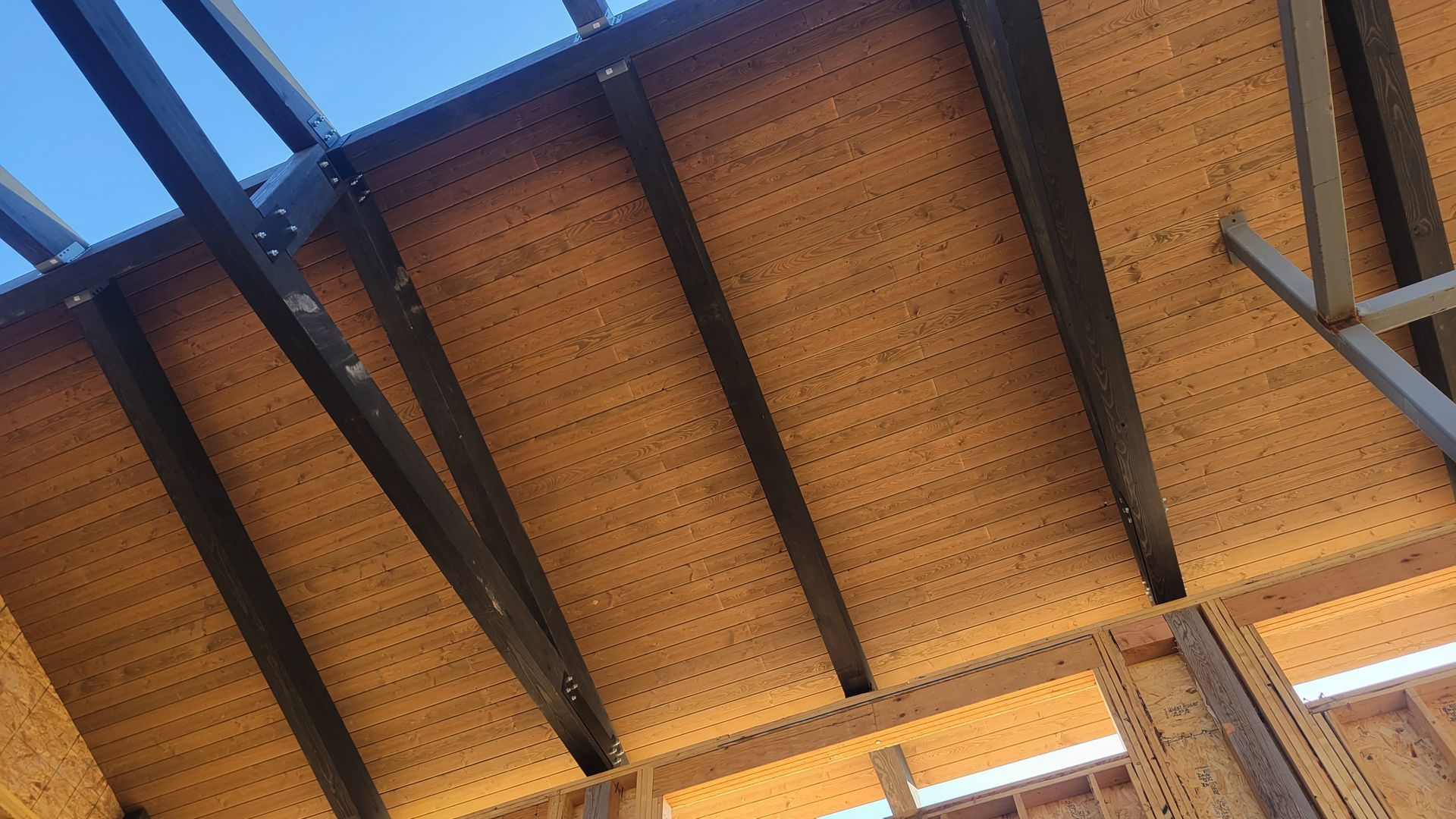 Wooden ceiling structure with dark beams and perforations, viewed from below under blue sky.