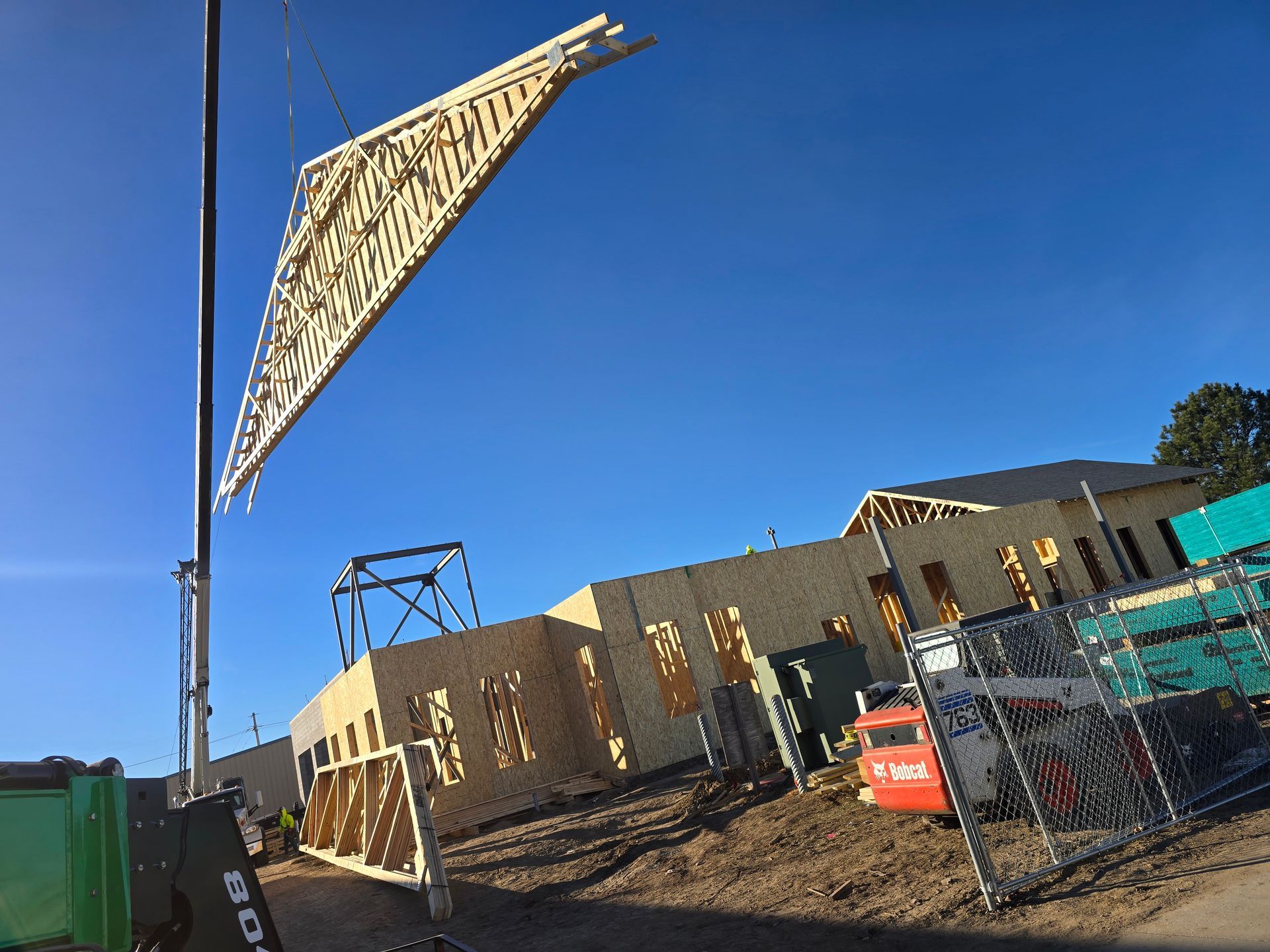 Crane lifting a wooden roof truss onto a building under construction, against a clear blue sky.