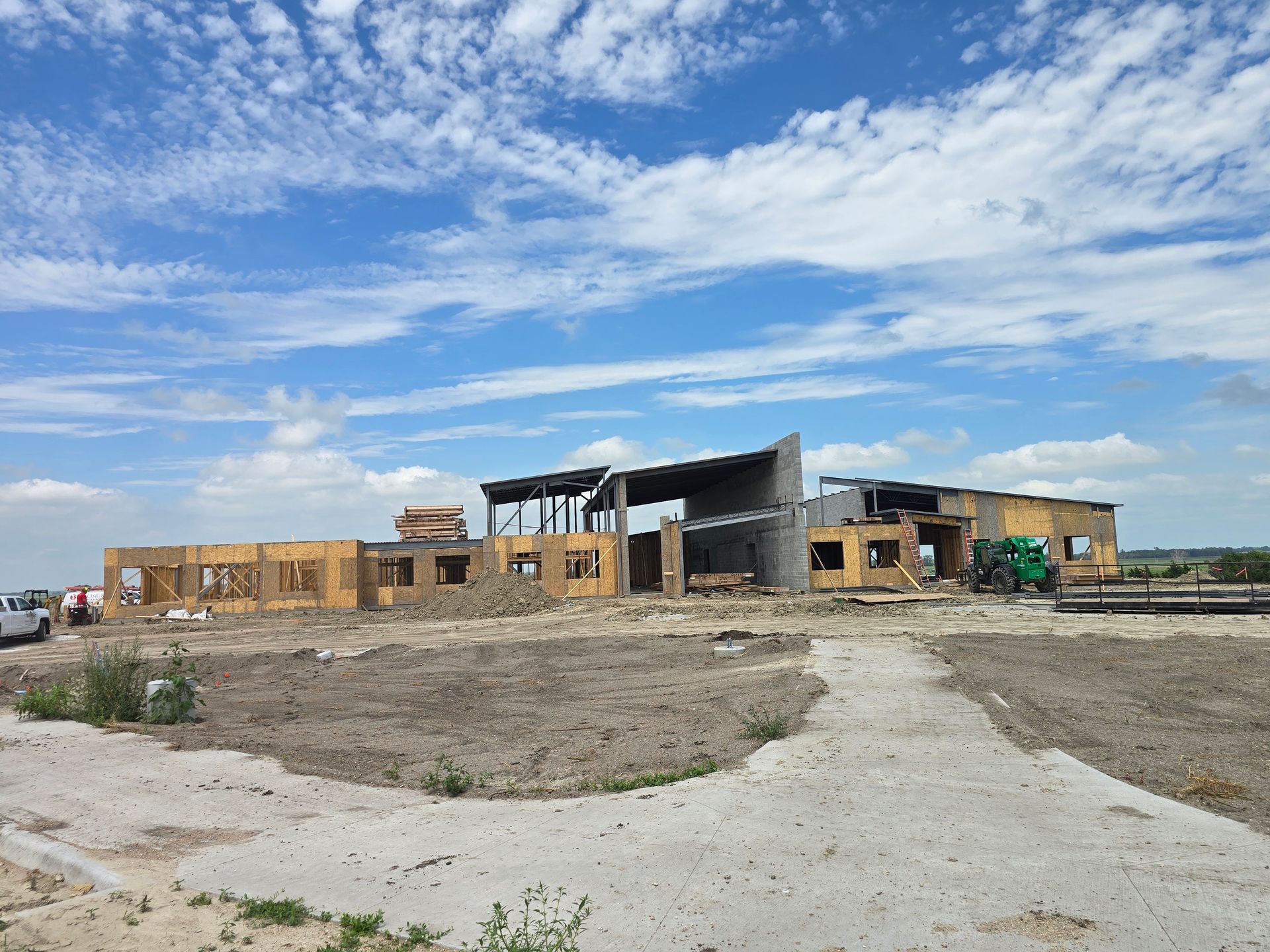 Construction site of a building under a blue sky with clouds; unfinished walls, concrete path in foreground.