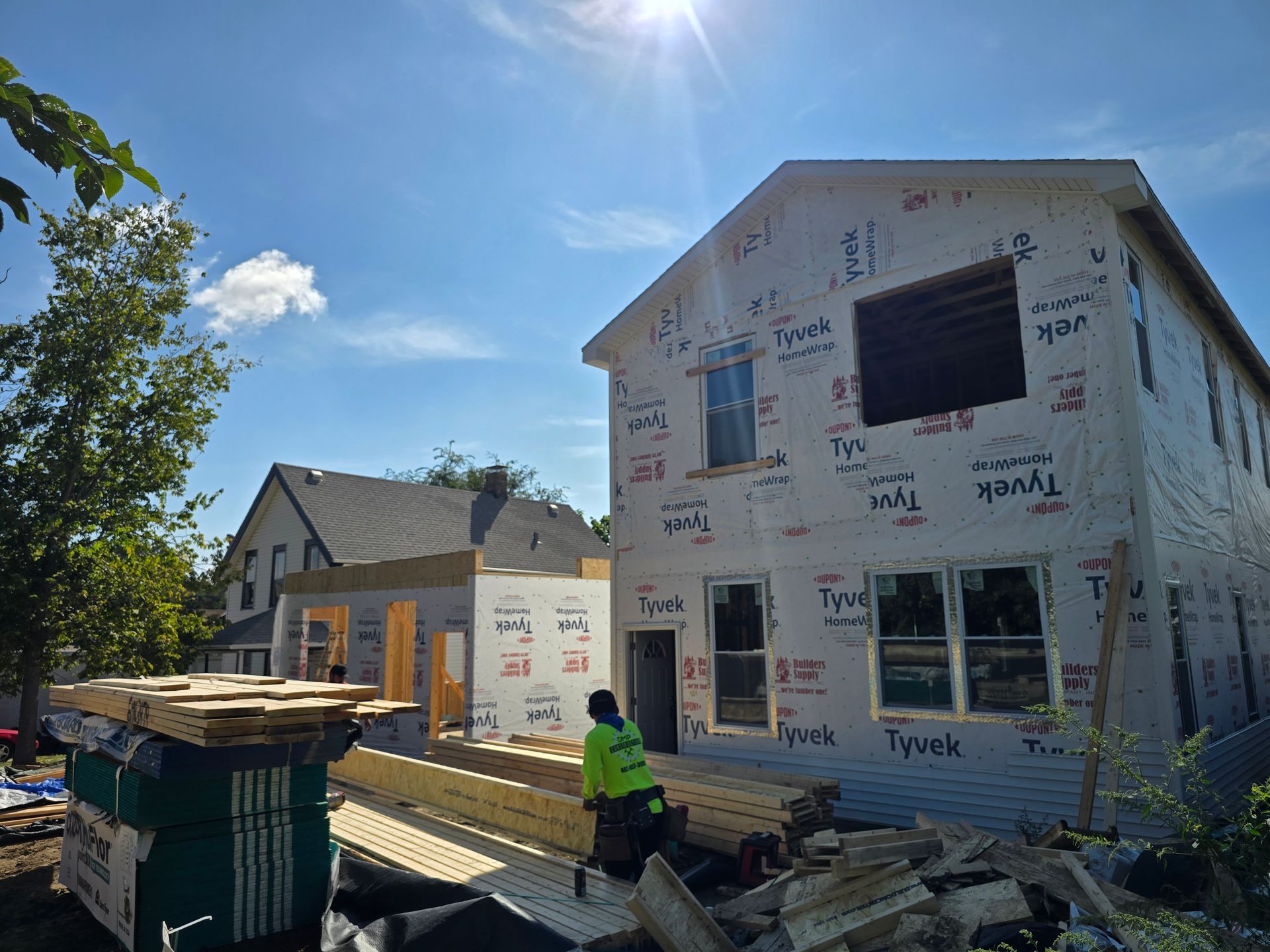 Two-story house under construction, wrapped in Tyvek. Sunlight, worker in green vest. Lumber and materials on site.