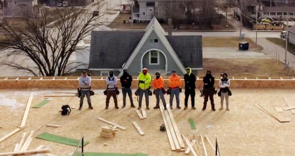 Construction workers stand on a roof, tools at their belts, a house in the background.