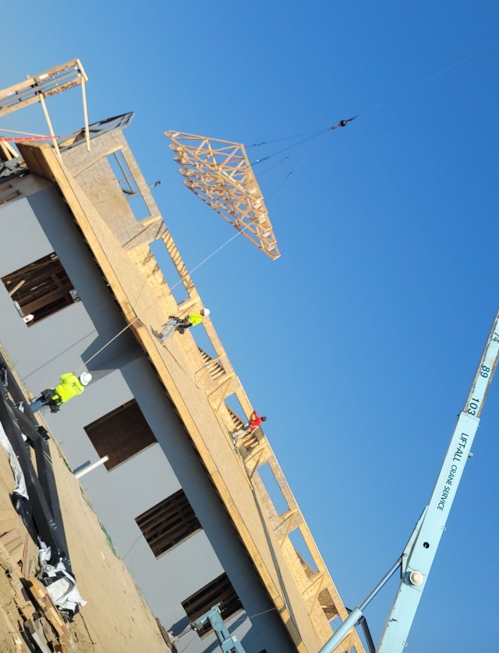 Construction workers lift a wooden truss with a crane onto a building frame against a blue sky.