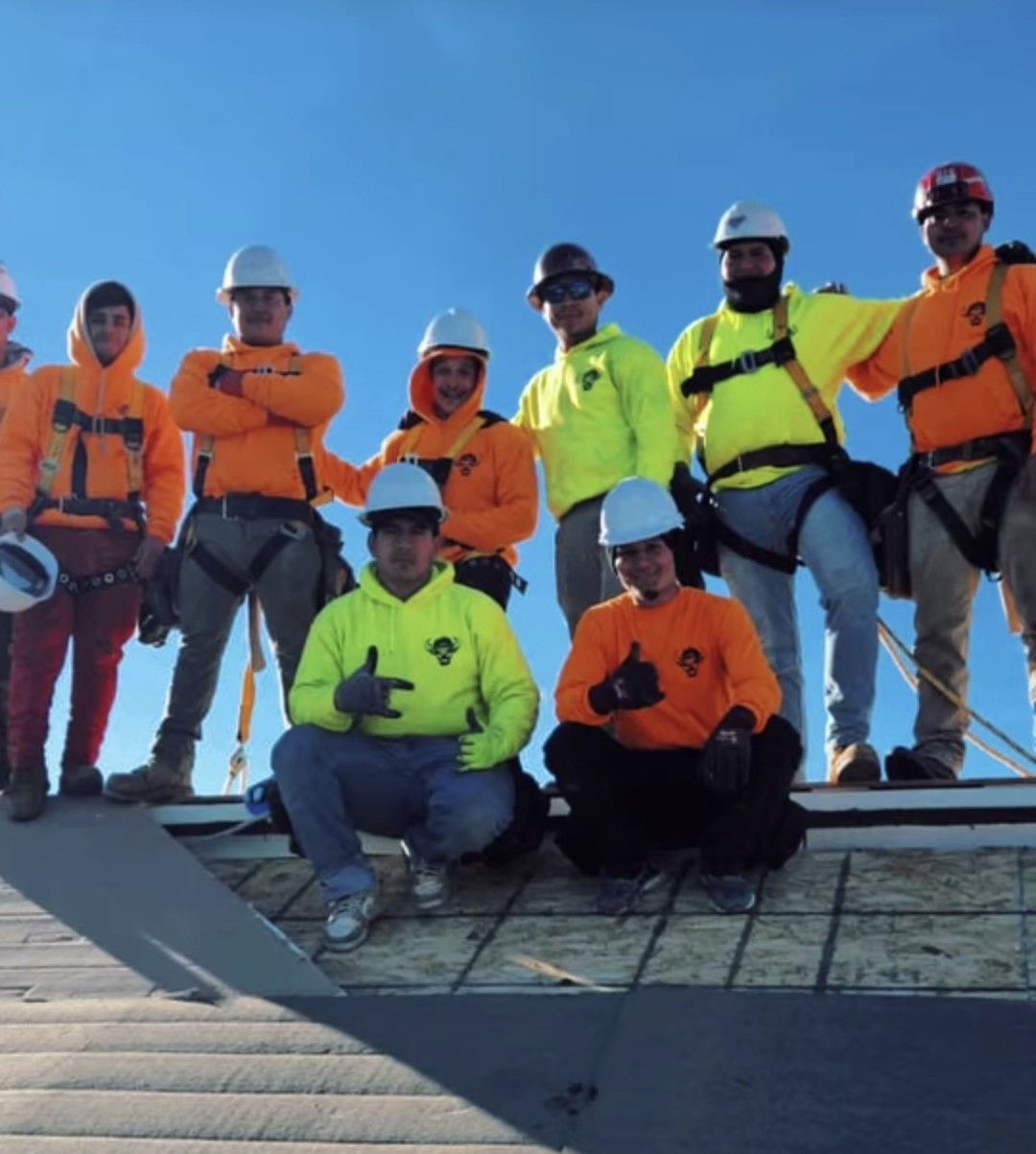 Group of construction workers on a rooftop posing for a photo, wearing safety gear and orange and yellow clothing.