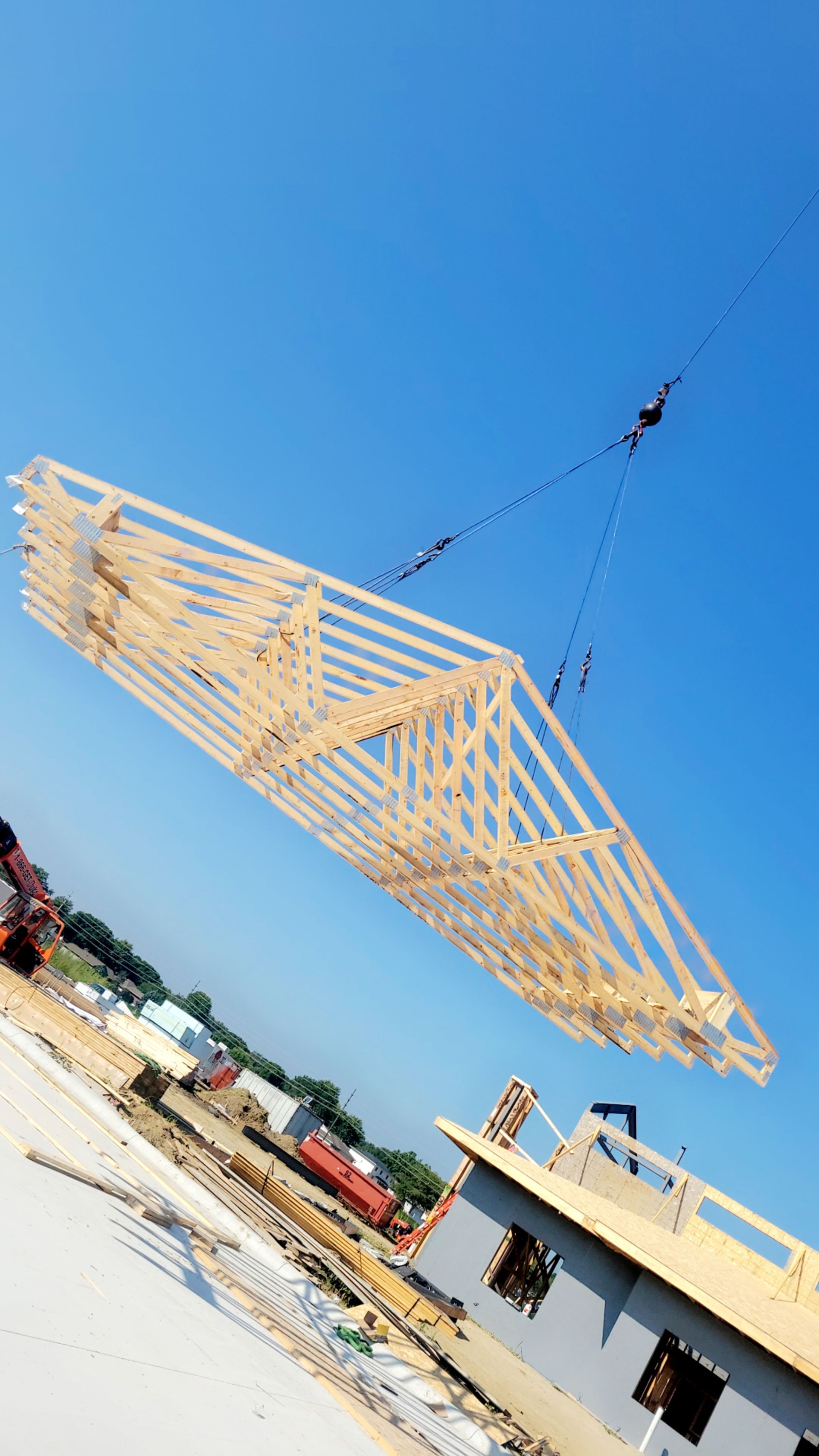 Crane lifting a large wooden roof truss against a clear blue sky at a construction site.