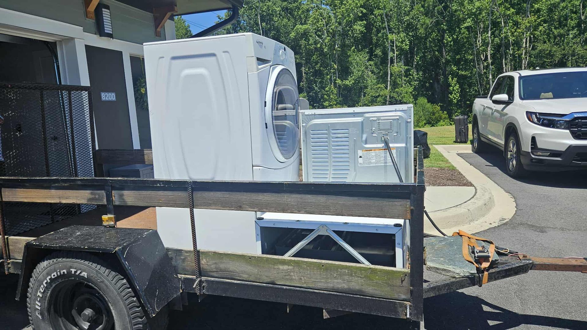 White appliances strapped on a trailer beside a driveway, with a white truck parked nearby.