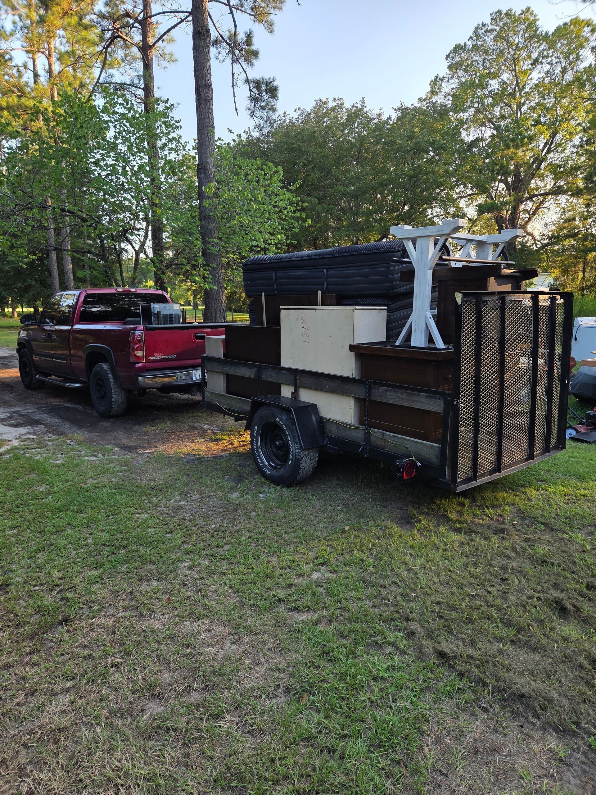Red pickup truck towing a black utility trailer loaded with stacked supplies in a grassy wooded area.
