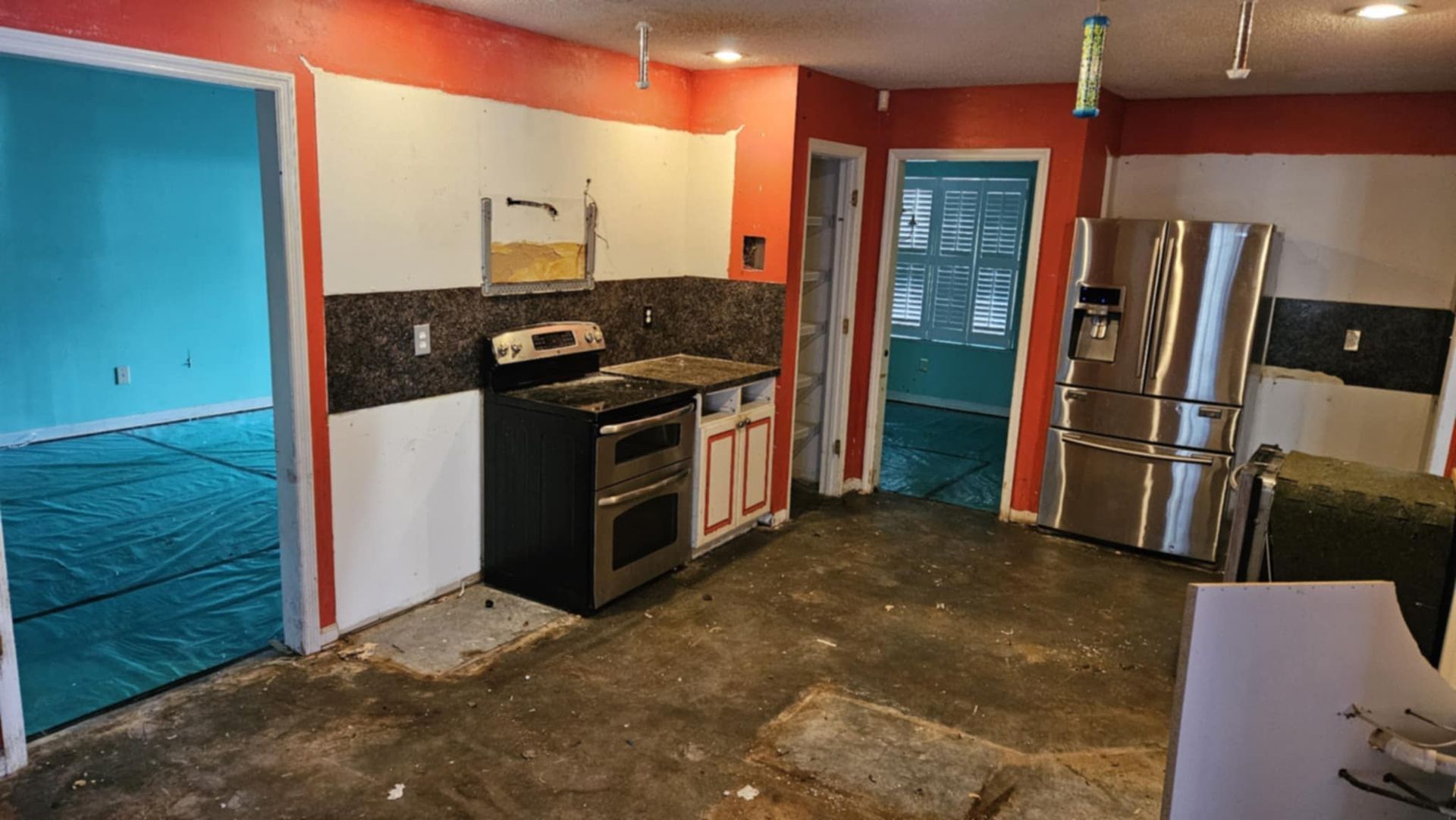 Damaged kitchen with orange walls, stainless fridge, stove, and debris-covered floor, likely under renovation