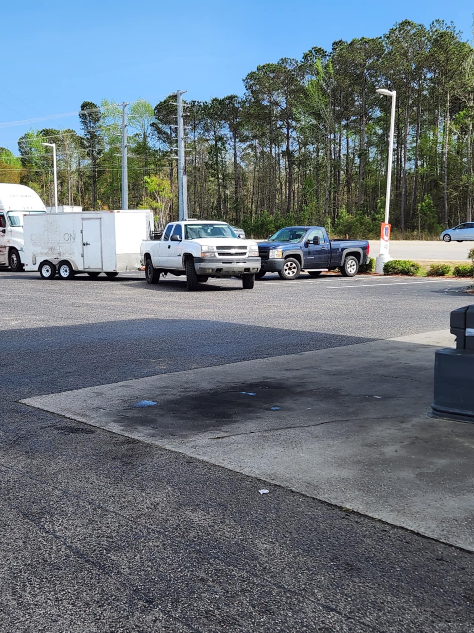 Parking lot with white trailer, pickup trucks, gravel ground, and trees in the background