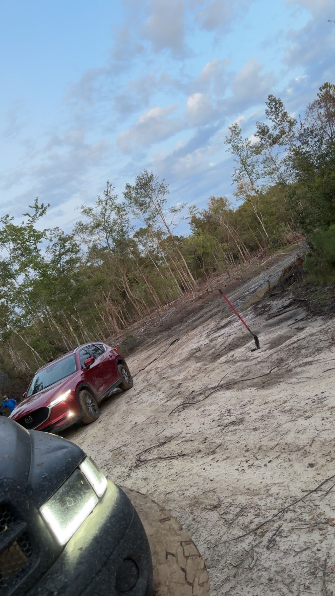 Two cars on a sandy dirt road beside sparse trees under a partly cloudy sky
