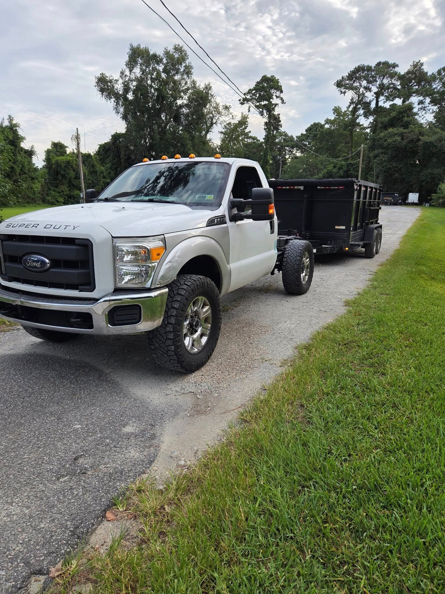 Silver Ford pickup towing a black dump trailer on a gravel road beside green grass and trees