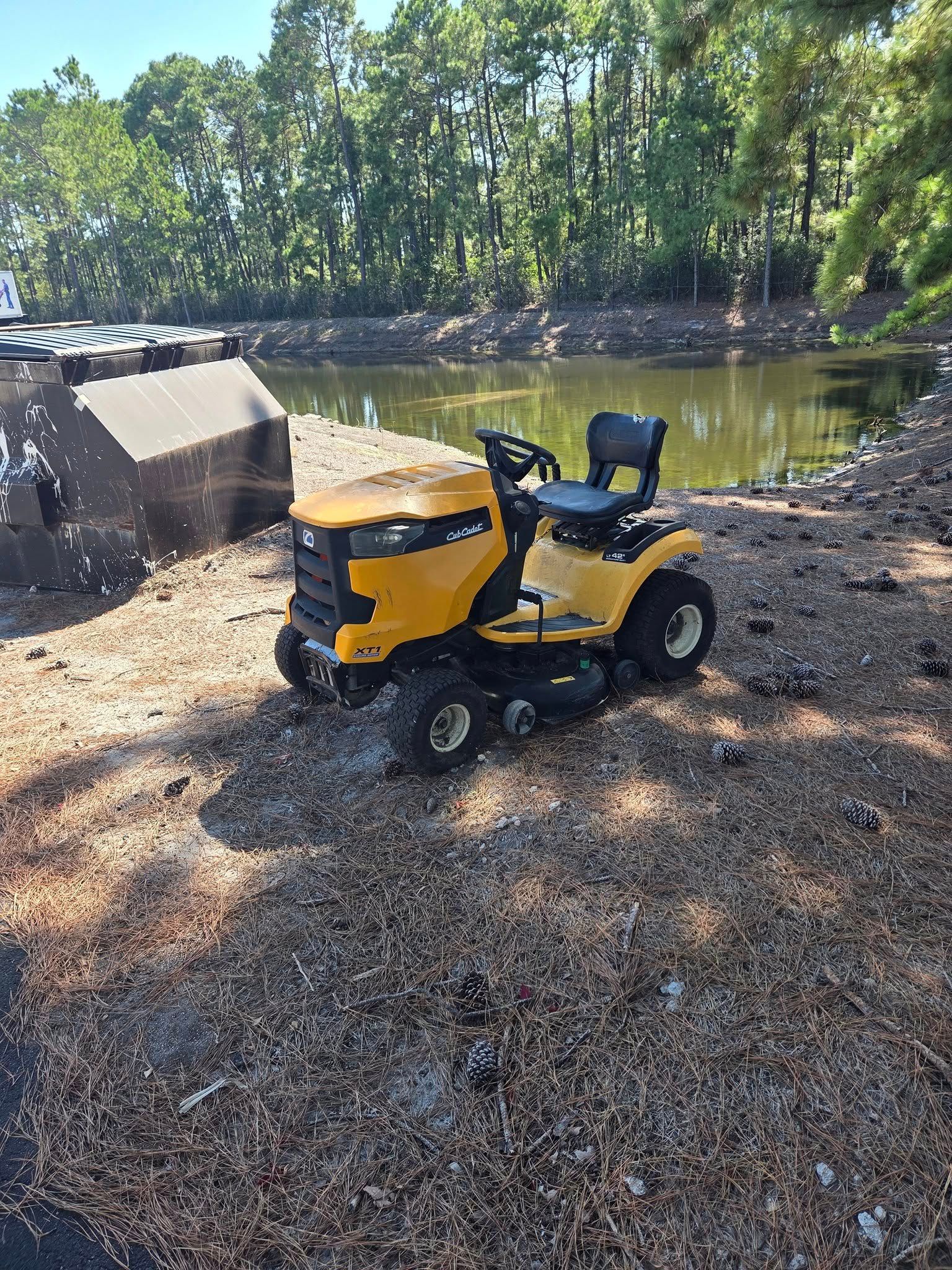 Yellow and black lawn tractor parked on a gravelly shoreline beside a calm river and trees.