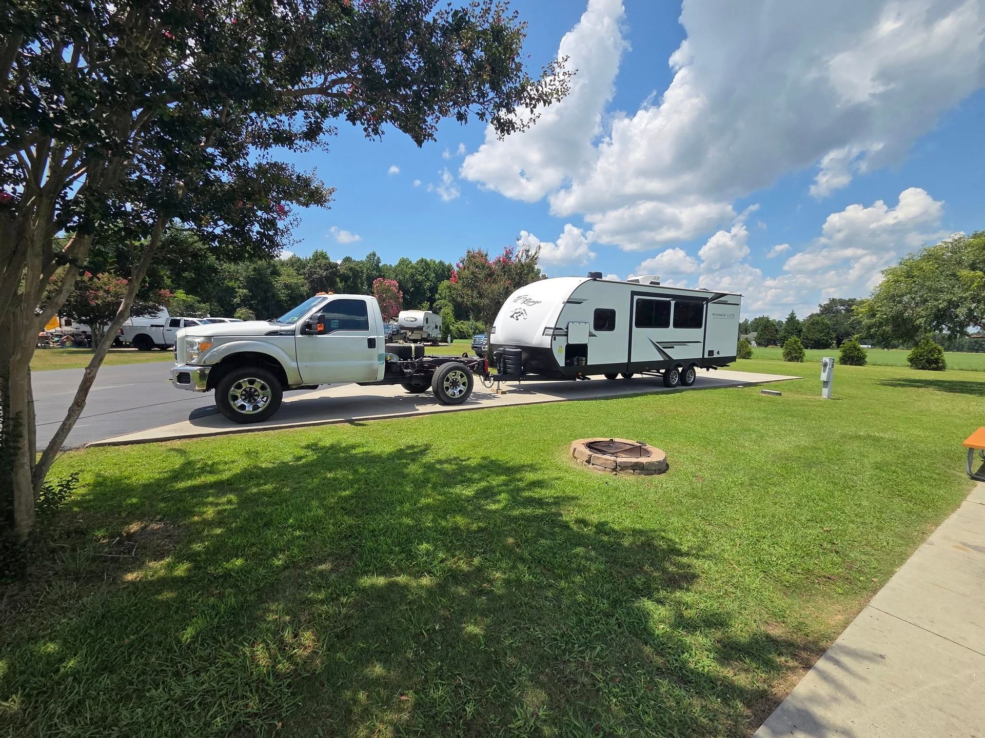 White pickup towing a small camper beside a grassy park under a blue sky