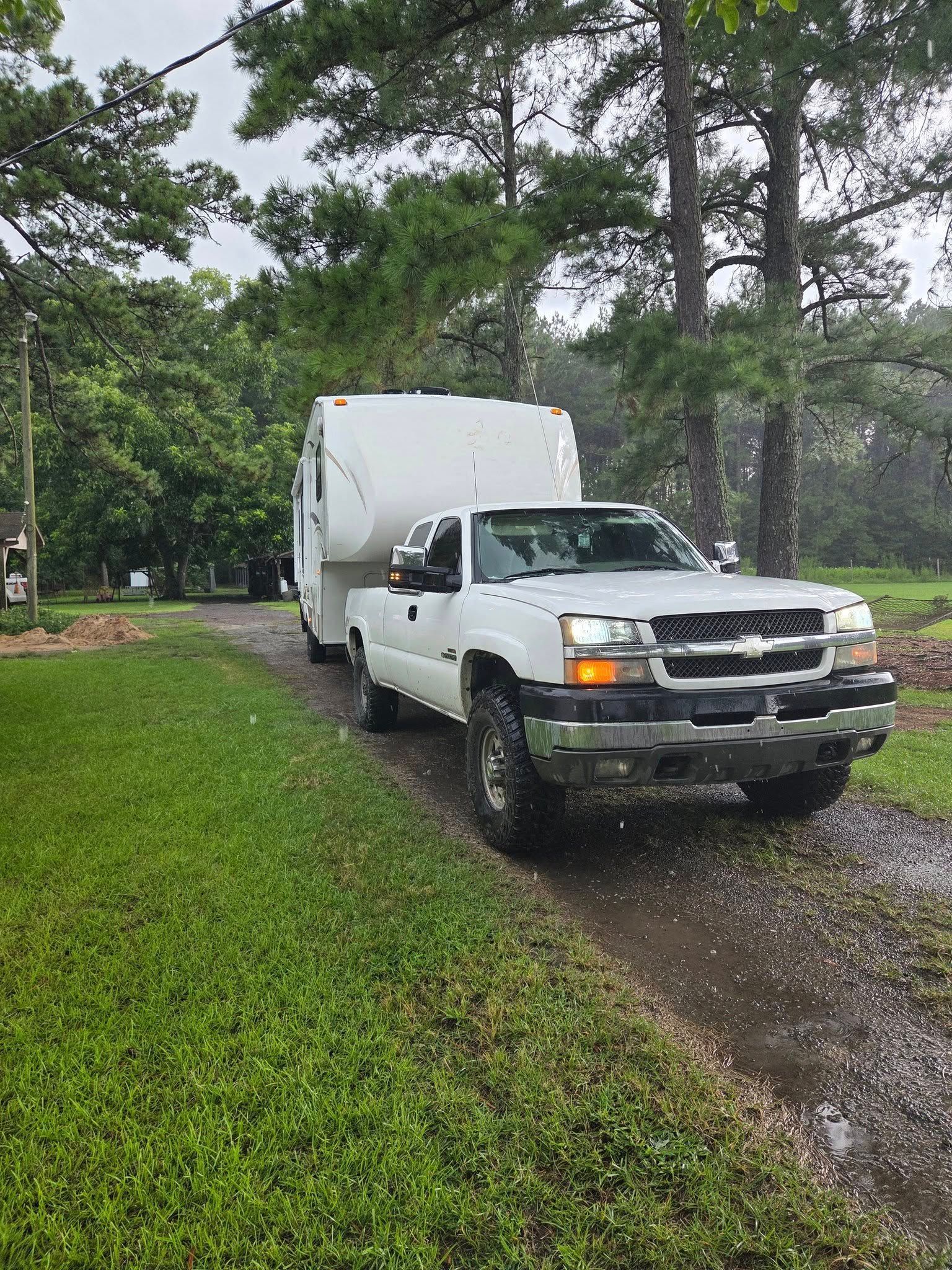 White box truck on a wet gravel driveway beside a grassy yard and tall trees